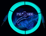 The image shows a coiled 10-foot Pig Hog instrument cable glowing bright cyan/turquoise against a dark background. It features glow-in-the-dark properties, standard 1/4" plugs on both ends, and the Pig Hog cartoon pig logo. Designed for electric guitars or basses, this vintage-style cable combines functionality with a unique glow effect, ideal for low-light stage performances.