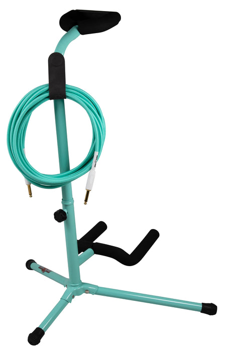 The image shows a seafoam green guitar stand with black padding and accents, featuring a tripod base with adjustable legs and rubber feet. It has an adjustable neck cradle and fold-out body supports, all padded to protect the guitar. A matching coiled turquoise cable with gold 1/4" plugs is neatly stored around the vertical post. This practical stand combines instrument support and cable management in one adjustable, space-saving design.