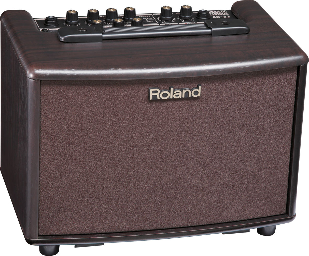 The image shows a Roland AC-33 acoustic guitar amplifier with a brown/rosewood finish and textured speaker grille. It features multiple control knobs for volume, EQ, and effects, including chorus, as indicated on the panel. The compact amp bears classic Roland branding and is designed to amplify acoustic guitars with clear, natural sound and built-in effects like chorus and reverb.