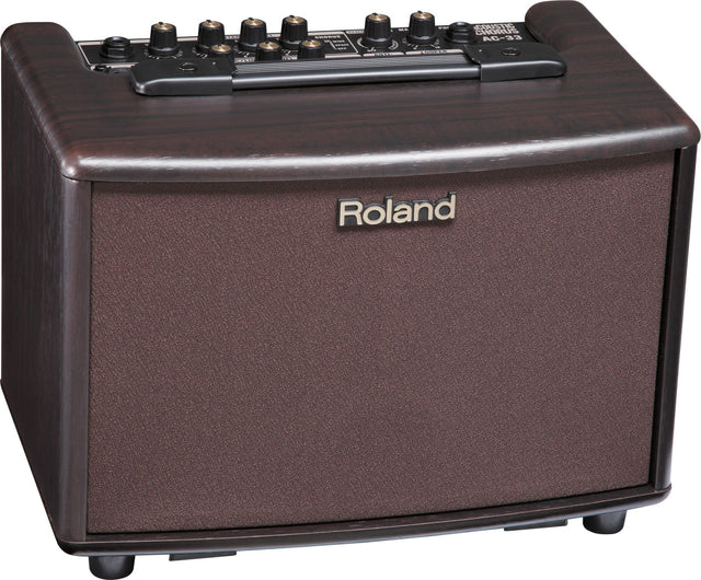 The image shows a Roland AC-33 acoustic guitar amplifier with a brown/rosewood finish and textured speaker grille. It features multiple control knobs for volume, EQ, and effects, including chorus, as indicated on the panel. The compact amp bears classic Roland branding and is designed to amplify acoustic guitars with clear, natural sound and built-in effects like chorus and reverb.