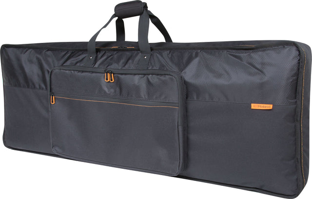 The image shows a Roland keyboard gig bag, a large, professional soft case for protecting and transporting keyboards. It has a sleek black design with orange accents, the Roland logo, padded comfort handles, multiple exterior pockets for accessories, and is made from durable nylon material. The long, rectangular shape is typical for keyboard cases, ideal for musicians on the move.