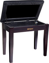 The image shows a Roland piano bench with a dark wood finish and gold Roland logo. It features a padded black seat that lifts to reveal a fabric-lined storage compartment. The bench has sturdy wooden legs and an elegant, professional design, suitable for home or studio use. It offers adjustable height for comfortable playing and convenient storage for sheet music and accessories.