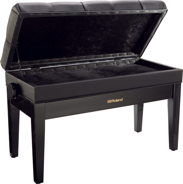 The image shows a Roland piano bench with a sleek black finish, padded tufted seat, adjustable height knob, and an open storage compartment lined with black fabric beneath the seat. It has four sturdy legs and the Roland logo on the front. This bench offers comfortable seating and convenient storage for sheet music and accessories, making it a popular choice for piano and keyboard players.