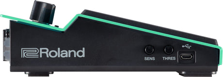 The image shows a black Roland electronic drum module with green accents, featuring two control knobs labeled "SENS" and "THRES," a USB port, and a trigger input jack. This module converts drum pad or trigger signals into electronic sounds, allowing sensitivity and threshold adjustments to optimize response. It connects to computers via USB for recording and integration with digital audio workstations.