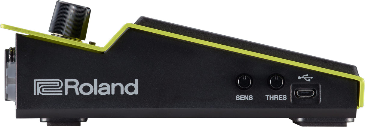 The image shows a Roland electronic drum trigger pad with a sleek black design and bright green/yellow trim. Visible features include the Roland logo, two control knobs labeled "SENS" and "THRES" for sensitivity and threshold adjustments, a USB port, and a trigger input jack. Its wedge shape is typical for drum modules, used by drummers to trigger electronic sounds with customizable response settings.