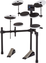 The image shows an electronic drum kit with multiple black and white drum pads, three cymbal pads, a digital sound module with an LCD display, an adjustable metal rack, two bass drum pedals, and connecting cables. It features velocity-sensitive pads, built-in sounds, volume control, headphone output, compact design, and MIDI connectivity. The Roland logo is visible on the sound module.