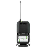 The image shows a Shure wireless bodypack transmitter with a black plastic housing and visible "SHURE" branding. It features an extendable antenna, an open battery compartment with two AA batteries, and a control panel with buttons and an LED indicator. This compact device wirelessly transmits audio from instruments or microphones, offering musicians freedom of movement during live performances.
