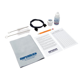 The image shows a Superslick brass instrument maintenance kit containing cleaning brushes, valve oil, a small container, a gray Superslick polishing cloth, instruction materials, a pencil, and a flexible pull-through cleaner. This kit provides essential tools for cleaning, lubricating, and maintaining brass instruments like trumpets and trombones to ensure optimal performance and sound quality.