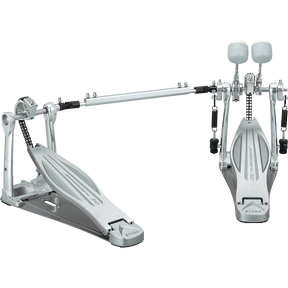 The image shows a high-end double bass drum pedal with dual foot pedals connected by a drive shaft, featuring chain or belt drive, adjustable white/felt beaters, sturdy metal frame, spring returns, and customizable tension and positioning. Made by Tama, this professional-grade pedal enables drummers to play fast, complex bass drum patterns used in metal, rock, and jazz.