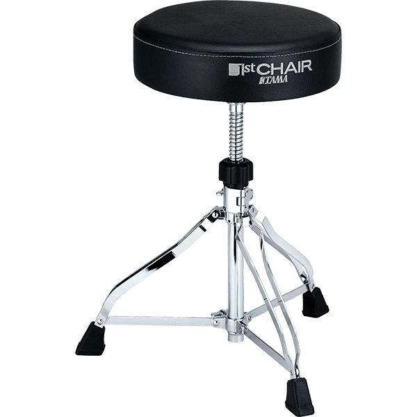 The image shows a Tama "1st Chair" drum throne with a round, padded black seat and visible branding. It features a height-adjustable threaded post, a chrome-plated tripod base with three legs, and black rubber feet for stability and floor protection. This professional-grade drum stool is essential for drummer comfort, posture, and performance.