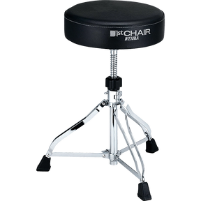 The image shows a Tama "1st Chair" drum throne with a round, padded black seat and visible branding. It features a height-adjustable threaded post, a chrome-plated tripod base with three legs, and black rubber feet for stability and floor protection. This professional-grade drum stool is essential for drummer comfort, posture, and performance.