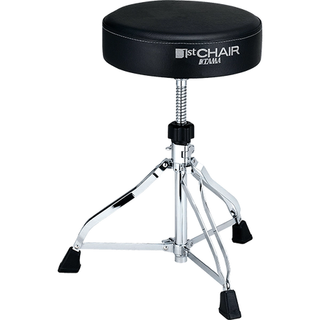 The image shows a Tama "1st Chair" drum throne with a round, padded black seat and visible branding. It features a height-adjustable threaded post, a chrome-plated tripod base with three legs, and black rubber feet for stability and floor protection. This professional-grade drum stool is essential for drummer comfort, posture, and performance.