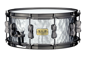 The image shows a TAMA S.L.P. snare drum with a distinctive hammered metal shell and black hardware. It features tension rods around the circumference, a snare throw-off lever, and the TAMA S.L.P. badge. This professional-quality drum, likely made of steel or aluminum, combines a textured finish that enhances both its look and sound, suitable for various musical styles.