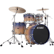 The image shows a complete TAMA drum kit featuring a bass drum with the logo, multiple tom-toms, a snare drum, and two cymbals. It has chrome hardware, including cymbal stands and adjustable supports. The drum shells display a gradient finish transitioning from light wood tones to dark blue-purple. This professional-quality set is suitable for live, studio, or practice use across various music styles.
