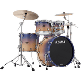 The image shows a complete TAMA drum kit featuring a bass drum with the logo, multiple tom-toms, a snare drum, and two cymbals. It has chrome hardware, including cymbal stands and adjustable supports. The drum shells display a gradient finish transitioning from light wood tones to dark blue-purple. This professional-quality set is suitable for live, studio, or practice use across various music styles.
