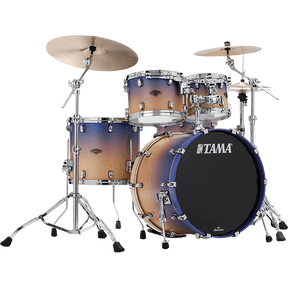 The image shows a complete TAMA drum kit featuring a bass drum with the logo, multiple tom-toms, a snare drum, and two cymbals. It has chrome hardware, including cymbal stands and adjustable supports. The drum shells display a gradient finish transitioning from light wood tones to dark blue-purple. This professional-quality set is suitable for live, studio, or practice use across various music styles.
