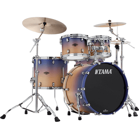 The image shows a complete TAMA drum kit featuring a bass drum with the logo, multiple tom-toms, a snare drum, and two cymbals. It has chrome hardware, including cymbal stands and adjustable supports. The drum shells display a gradient finish transitioning from light wood tones to dark blue-purple. This professional-quality set is suitable for live, studio, or practice use across various music styles.