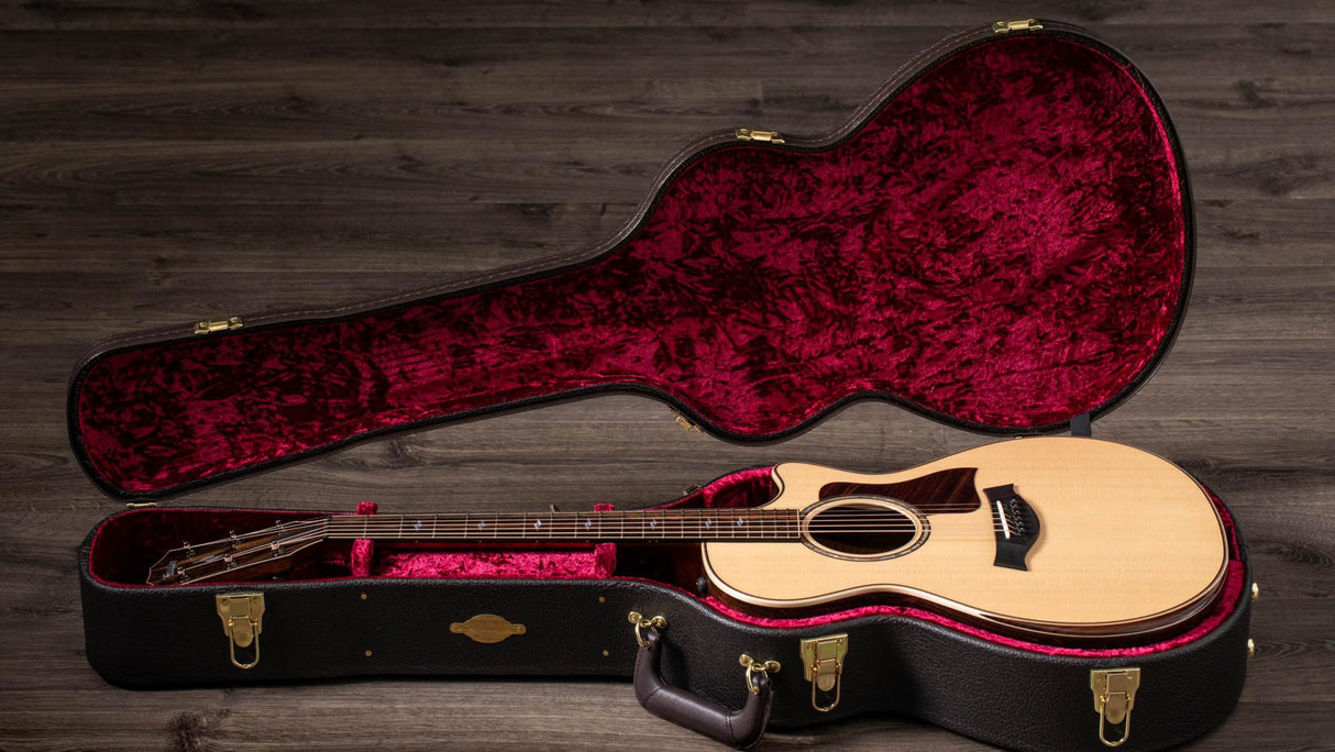 The image shows a natural wood acoustic guitar with steel strings and a cutaway body inside a black hard case. The case features brass hardware and a plush burgundy velvet interior molded to fit the guitar, with extra space for accessories and a handle. The setup rests on weathered wooden flooring, photographed with professional lighting, highlighting a premium instrument and protective case designed for serious musicians or collectors.