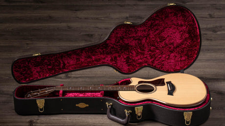 The image shows a natural wood acoustic guitar with steel strings and a cutaway body inside a black hard case. The case features brass hardware and a plush burgundy velvet interior molded to fit the guitar, with extra space for accessories and a handle. The setup rests on weathered wooden flooring, photographed with professional lighting, highlighting a premium instrument and protective case designed for serious musicians or collectors.
