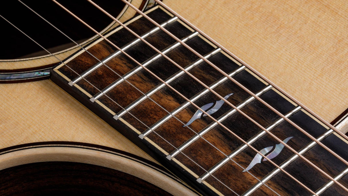The image shows a close-up of an acoustic guitar focusing on the sound hole, fretboard, and strings. Visible elements include the circular sound hole with decorative rosette, six steel strings, metal frets, dark wood fretboard with position markers, and light-colored soundboard. The guitar features clean craftsmanship and natural wood tones, reflecting a well-made, versatile instrument suited for various musical styles.