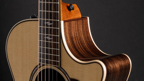 The image shows a beautifully crafted acoustic-electric guitar with a light-colored spruce top and dark rosewood back and sides, featuring elegant binding and a cutaway design. It has a dark fretboard with decorative inlays, quality tuning pegs, and a visible output jack. Captured with professional lighting against a dark background, it highlights fine craftsmanship and premium materials typical of a high-end, handcrafted instrument.