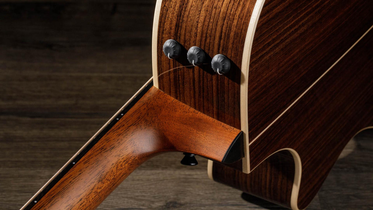 The image shows a close-up of an acoustic-electric guitar’s lower bout and side, featuring rich, dark wood with lighter binding. Three control knobs for the built-in pickup or preamp system are visible, highlighting the guitar’s ability to be amplified. The craftsmanship showcases detailed wood grain and finishing. The guitar rests on a wooden surface, combining natural beauty with modern electronics.