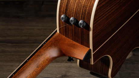 The image shows a close-up of an acoustic-electric guitar’s lower bout and side, featuring rich, dark wood with lighter binding. Three control knobs for the built-in pickup or preamp system are visible, highlighting the guitar’s ability to be amplified. The craftsmanship showcases detailed wood grain and finishing. The guitar rests on a wooden surface, combining natural beauty with modern electronics.