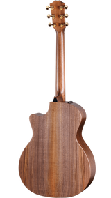 The image shows the back of an acoustic guitar with a polished medium to dark brown wood finish, likely mahogany. It features a traditional body shape with a cutaway for easier fret access. Six gold-colored tuning pegs are visible on the headstock. A small black component, likely an output jack, suggests it’s an acoustic-electric model. The guitar displays clean lines, well-executed binding, and professional craftsmanship.