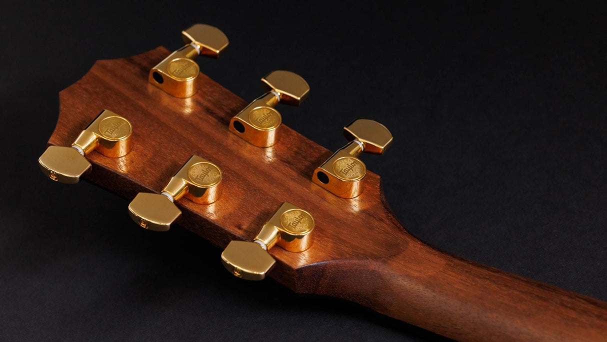 The image shows the headstock of a high-quality acoustic guitar featuring rich reddish-brown wood grain, gold-plated sealed gear tuning machines, and professional hardware. The tuning pegs have embossed brand markings, indicating a premium instrument. These components reflect careful craftsmanship and are essential for tuning the guitar’s strings accurately.