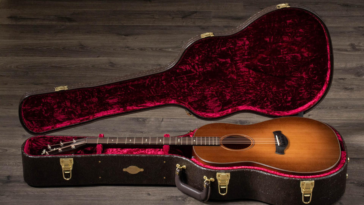 The image shows a honey-colored acoustic guitar with a classical body, dark fretboard, and visible tuning pegs inside a black hard case. The case features a plush deep red interior, gold latches, and a form-fitted design. It rests open on a dark wooden floor, highlighting the guitar’s craftsmanship and suggesting a professional-quality instrument in a premium protective case.