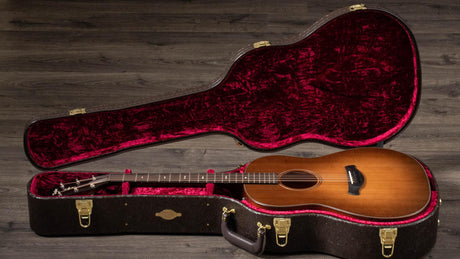 The image shows a honey-colored acoustic guitar with a classical body, dark fretboard, and visible tuning pegs inside a black hard case. The case features a plush deep red interior, gold latches, and a form-fitted design. It rests open on a dark wooden floor, highlighting the guitar’s craftsmanship and suggesting a professional-quality instrument in a premium protective case.