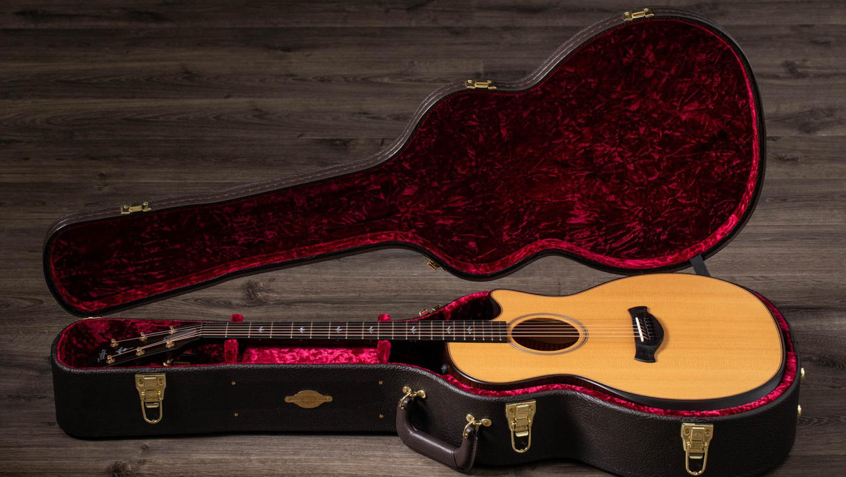 The image shows a natural wood-finished acoustic guitar with a light spruce top, dark back and sides, and a dark fretboard inside an open black hard shell case. The case features deep red plush lining and gold-colored hardware. Both rest on weathered wooden flooring, captured in professional lighting, highlighting a high-quality instrument and protective case designed for serious musicians.