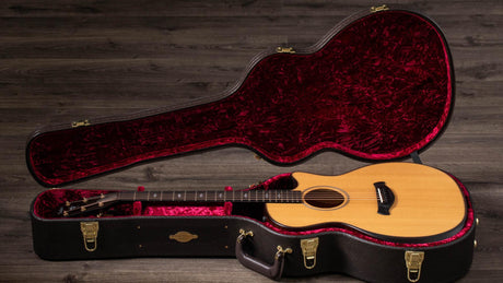 The image shows a natural wood-finished acoustic guitar with a light spruce top, dark back and sides, and a dark fretboard inside an open black hard shell case. The case features deep red plush lining and gold-colored hardware. Both rest on weathered wooden flooring, captured in professional lighting, highlighting a high-quality instrument and protective case designed for serious musicians.