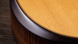 The image shows a close-up of an acoustic guitar’s body, featuring the dark binding around the soundhole, a light-colored spruce soundboard with a distinctive cross-grain pattern, and a darker hardwood side, possibly rosewood or mahogany. The contrast between the woods and the clean binding highlights the guitar’s quality craftsmanship and careful wood selection.