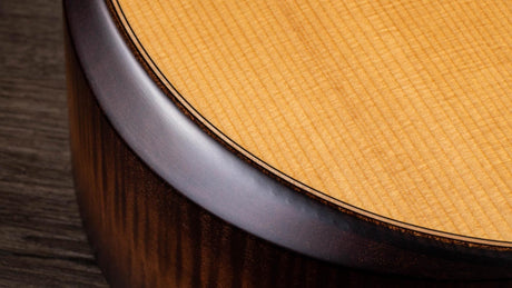 The image shows a close-up of an acoustic guitar’s body, featuring the dark binding around the soundhole, a light-colored spruce soundboard with a distinctive cross-grain pattern, and a darker hardwood side, possibly rosewood or mahogany. The contrast between the woods and the clean binding highlights the guitar’s quality craftsmanship and careful wood selection.