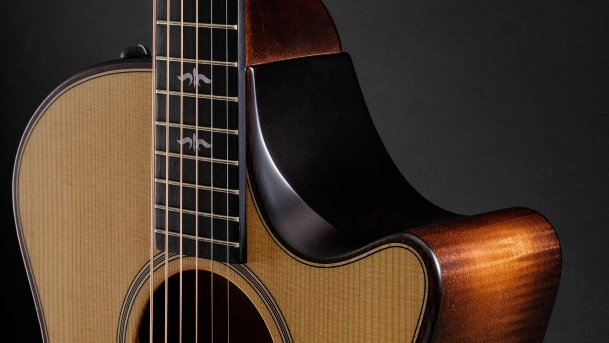 The image shows a high-quality acoustic guitar with a cutaway body for easy high-fret access. It features a natural wood finish with visible grain, dark binding around the edges, steel strings, metal frets on the fretboard, tuning pegs on the headstock, and a partially visible sound hole. The guitar has a glossy finish and refined craftsmanship, making it suitable for various musical styles.