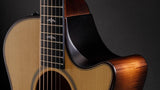 The image shows a high-quality acoustic guitar with a cutaway body for easy high-fret access. It features a natural wood finish with visible grain, dark binding around the edges, steel strings, metal frets on the fretboard, tuning pegs on the headstock, and a partially visible sound hole. The guitar has a glossy finish and refined craftsmanship, making it suitable for various musical styles.