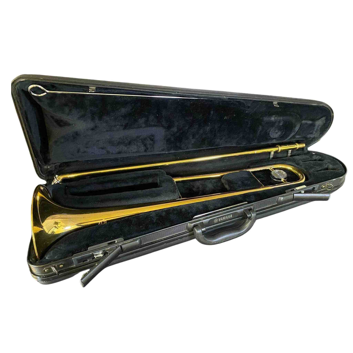 The image shows a Yamaha brass trombone in its black hard case with a dark velvet interior. The trombone features the classic golden finish and slide mechanism. The case includes combination locks and a handle. This professional-quality instrument is used in orchestras, jazz bands, and more, producing sound by buzzing lips in the mouthpiece and changing pitch with the slide.